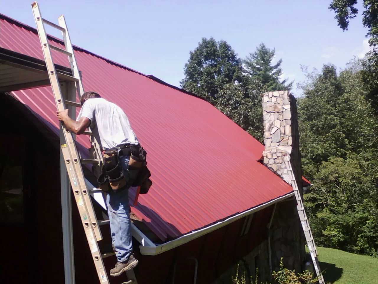 Roofer on a ladder beside a red metal roof and stone chimney — Soffit & Fascia Repair by Roofing Brothers in Buncombe
