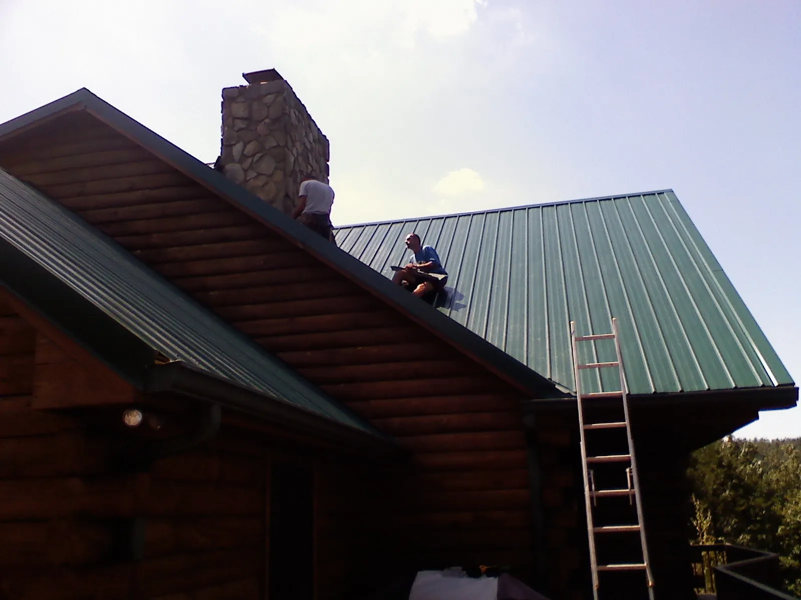 Roofers installing green metal panels beside a stone chimney on a log home in Buncombe